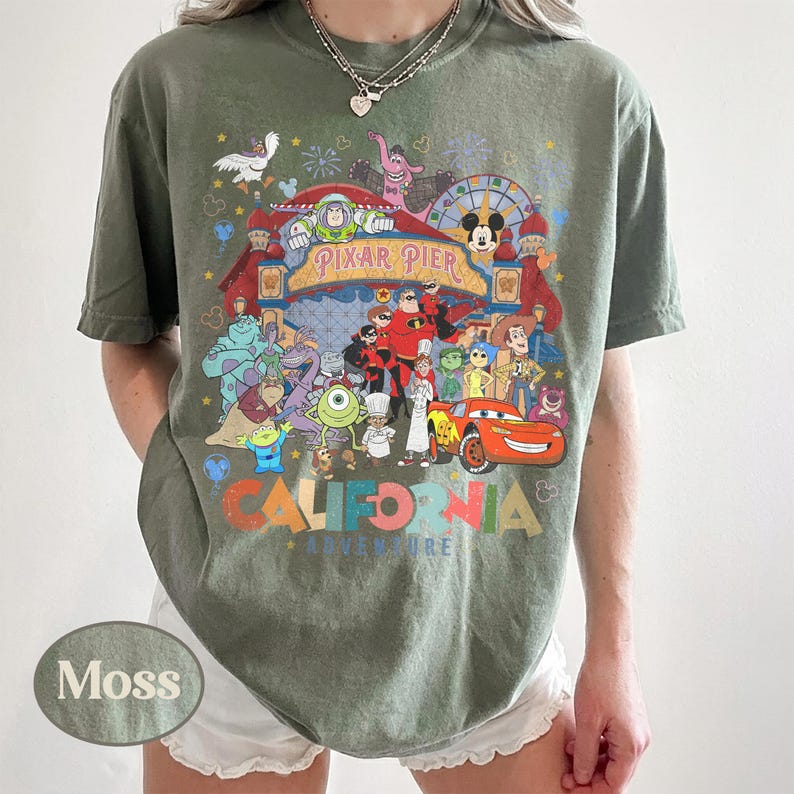 May include: Moss green t-shirt featuring a colorful graphic of Pixar Pier characters and the text "CALIFORNIA ADVENTURE". The shirt has a crew neck and short sleeves. The shirt is a relaxed fit.