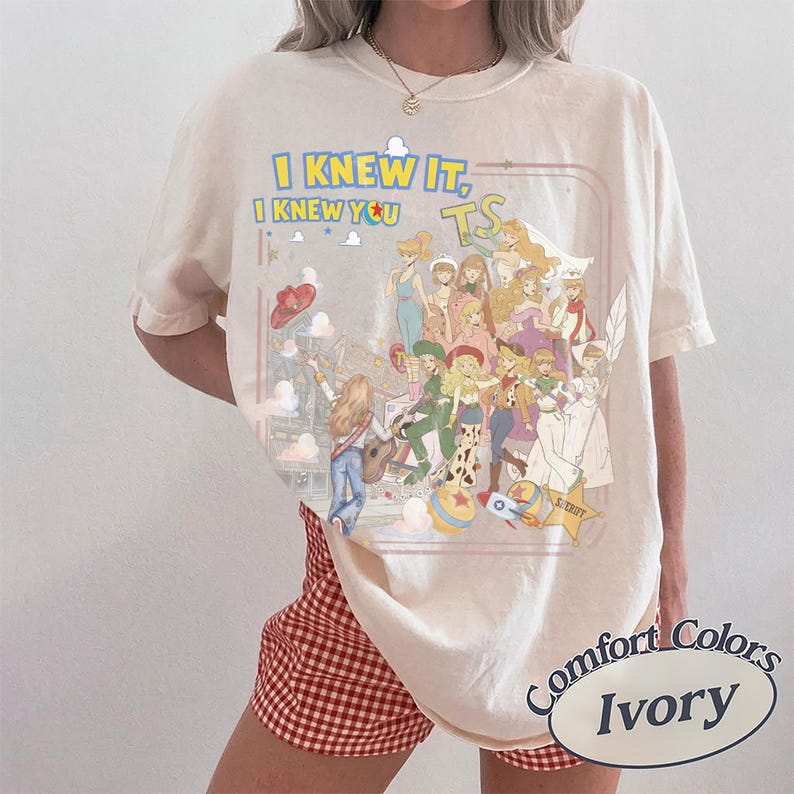 May include: An ivory short-sleeve t-shirt featuring a graphic print with the text I KNEW IT, I KNEW YOU TS. The design displays a collage of various animated characters in colorful outfits, including a cowboy, a space ranger, and a sheriff star.