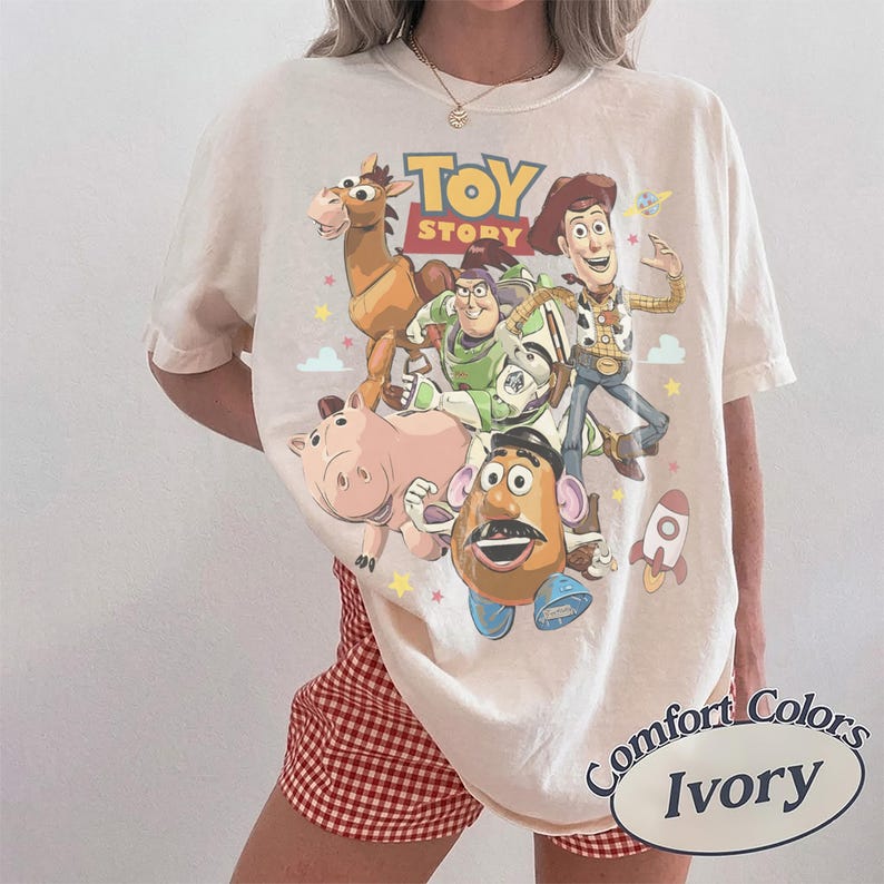 May include: An ivory short-sleeve t-shirt featuring a colorful graphic print of Toy Story characters including Woody, Buzz Lightyear, Bullseye, Hamm, and Mr. Potato Head, with the text "TOY STORY" printed in bold yellow and red letters.