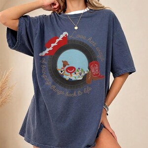 May include: A dark blue short-sleeve t-shirt featuring a graphic print of a tire framing a scene with a cowboy doll, a space ranger doll, and a red cowboy boot. The text "Love has ways of bringing things back to life" is printed in a circular script.