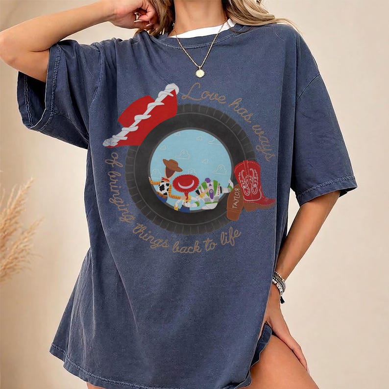 May include: A dark blue short-sleeve t-shirt featuring a graphic print of a tire framing a scene with a cowboy doll, a space ranger doll, and a red cowboy boot. The text "Love has ways of bringing things back to life" is printed in a circular script.