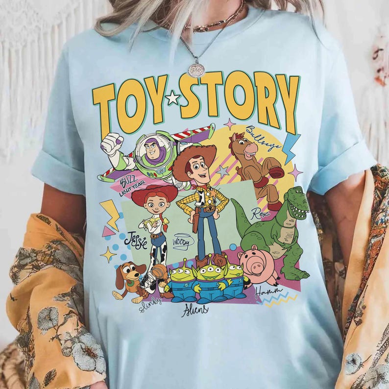 May include: Pale blue t-shirt with the words "TOY STORY" in yellow, and a star. The shirt features cartoon characters such as Buzz Lightyear, Woody, Jessie, and Rex. The t-shirt is a crew neck style.