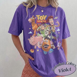 May include: A purple short-sleeve t-shirt featuring a graphic print of Toy Story characters including Woody, Buzz Lightyear, Bullseye, Hamm, and Mr. Potato Head. The text TOY STORY is printed in yellow above the characters, with small stars and clouds.