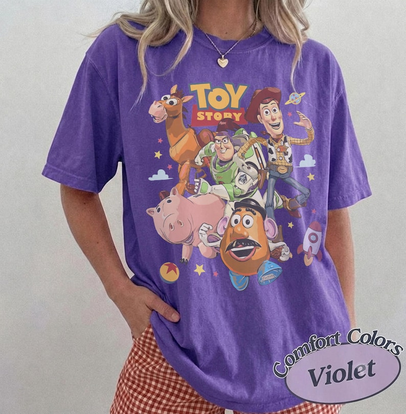 May include: A purple short-sleeve t-shirt featuring a graphic print of Toy Story characters including Woody, Buzz Lightyear, Bullseye, Hamm, and Mr. Potato Head. The text TOY STORY is printed in yellow above the characters, with small stars and clouds.