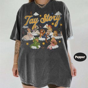 May include: A charcoal gray oversized t-shirt featuring the text Tay Story in yellow cursive font. The graphic includes multiple illustrated figures dressed as characters sitting on white clouds, surrounded by yellow stars and musical notes.