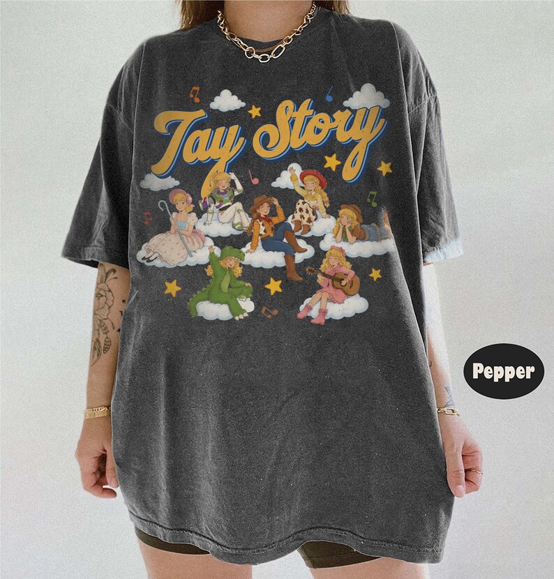 May include: A charcoal gray oversized t-shirt featuring the text Tay Story in yellow cursive font. The graphic includes multiple illustrated figures dressed as characters sitting on white clouds, surrounded by yellow stars and musical notes.