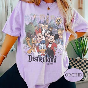 Puede incluir: Camiseta lila claro con un estampado de Disneyland que presenta a Mickey Mouse, Minnie Mouse, Donald Duck y Goofy. El estampado incluye el logotipo de Disneyland y "Est. 1955". La camiseta tiene mangas cortas y un corte holgado. La palabra "ORCHID" est&aacute; en una etiqueta.