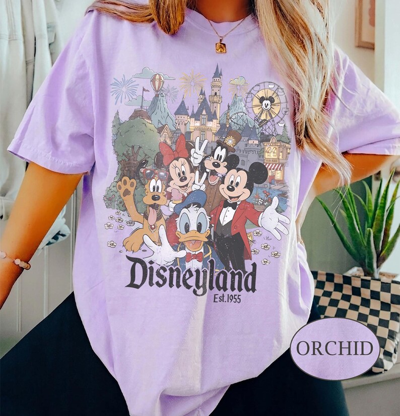Puede incluir: Camiseta lila claro con un estampado de Disneyland que presenta a Mickey Mouse, Minnie Mouse, Donald Duck y Goofy. El estampado incluye el logotipo de Disneyland y "Est. 1955". La camiseta tiene mangas cortas y un corte holgado. La palabra "ORCHID" est&aacute; en una etiqueta.
