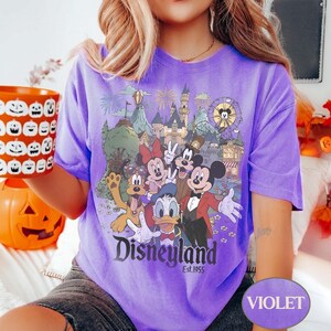 Puede incluir: Camiseta violeta con un dise&ntilde;o de Disneyland que presenta a Mickey Mouse, Minnie Mouse, Donald Duck y Pluto. El dise&ntilde;o incluye el logotipo de Disneyland y el texto "Est. 1955". La camiseta es de color violeta liso.
