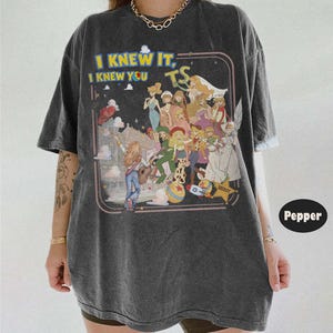 May include: A charcoal gray short-sleeve t-shirt featuring a graphic print with the text "I KNEW IT, I KNEW YOU TS" and an illustration of multiple animated characters in various outfits, including a sheriff star, a rocket, and a ball.