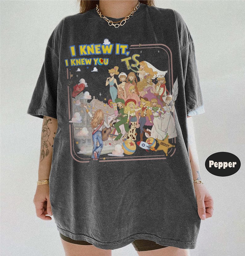 May include: A charcoal gray short-sleeve t-shirt featuring a graphic print with the text "I KNEW IT, I KNEW YOU TS" and an illustration of multiple animated characters in various outfits, including a sheriff star, a rocket, and a ball.