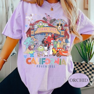 May include: Orchid-colored t-shirt featuring a colorful graphic of Pixar Pier characters and the words "California Adventure." The design includes characters from various Pixar films. The shirt has short sleeves.