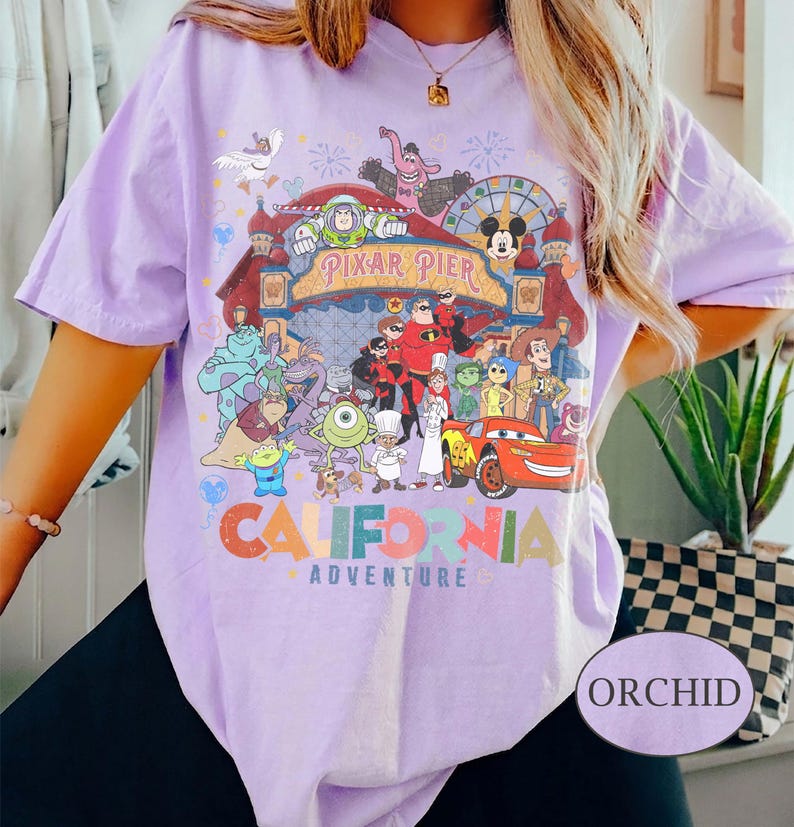 May include: Orchid-colored t-shirt featuring a colorful graphic of Pixar Pier characters and the words "California Adventure." The design includes characters from various Pixar films. The shirt has short sleeves.