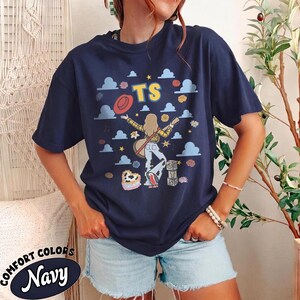 Op de afbeelding: Een marineblauw t-shirt met een grafische print van een persoon met lang haar die een gitaar vasthoudt, een hoed draagt en een broek met koeienvlekkenpatroon. Het ontwerp bevat de letters TS, blauwe wolken, sterren, bloemen en blokken.