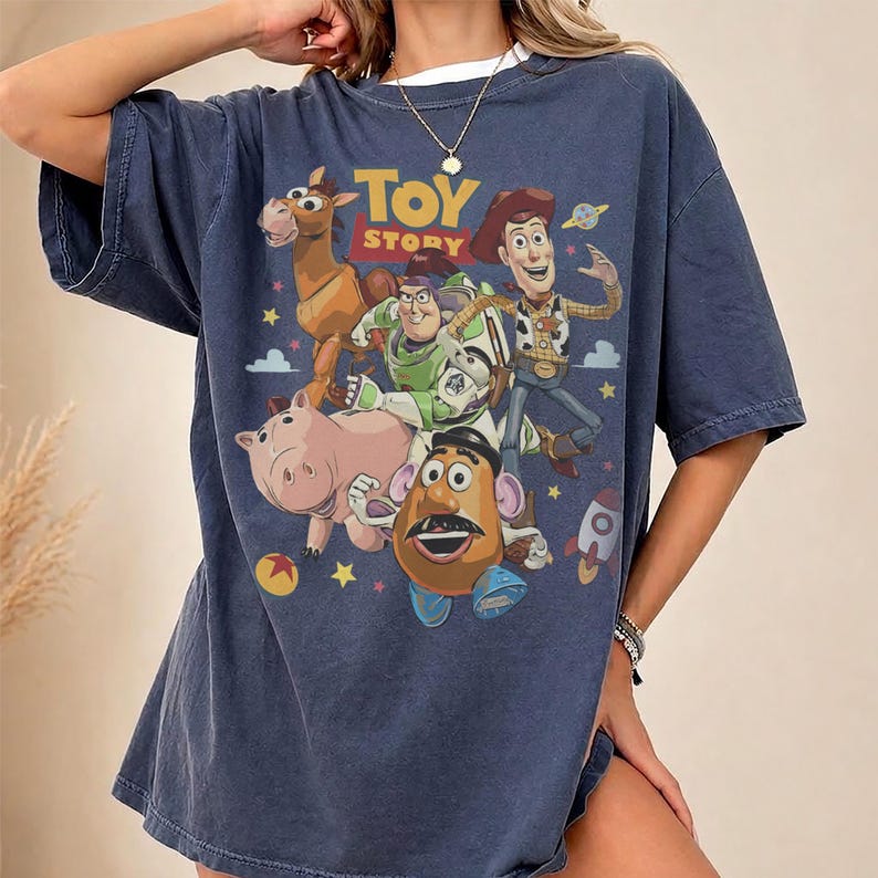 May include: A dark blue short-sleeve t-shirt featuring a graphic print of Toy Story characters including Woody, Buzz Lightyear, Bullseye, Hamm, and Mr. Potato Head. The text Toy Story is printed in yellow and red above the character group.