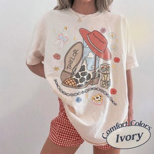 Puede incluir: Una camiseta de manga corta color marfil con un estampado gr&aacute;fico de una bota de vaquero con estampado de vaca con el nombre TAYLOR, un sombrero rojo, bloques apilados con las letras T, S y 13, un lazo, flores y el texto I KNEW YOU WERE TROUBLE.