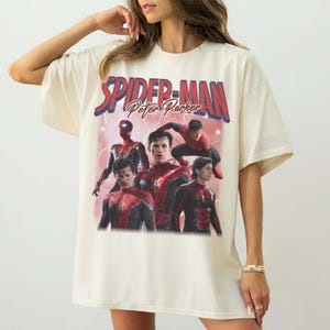 May include: A cream-coloured t-shirt with the text "SPIDER-MAN Peter Parker" in red and blue. The shirt features multiple images of Spider-Man in action poses. The design has a vintage aesthetic.