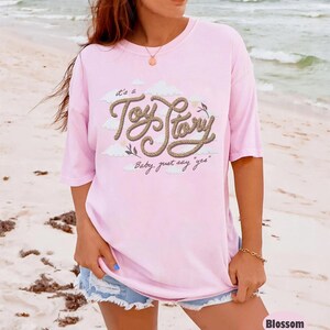 Pu&ograve; includere: Una t-shirt a maniche corte rosa chiaro con un design grafico che riporta il testo "it's a Toy Story" scritto con un carattere simile a una corda, accompagnato dalla frase "Baby, just say 'yes'" e illustrazioni di nuvole bianche e rametti.