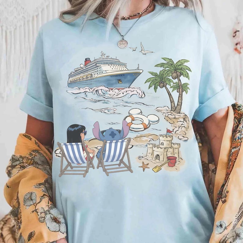 May include: Light blue t-shirt featuring a beach scene with a cruise ship, palm trees, and a sandcastle. The graphic includes cartoon characters Stitch and Lilo relaxing in beach chairs. A Mickey Mouse-shaped life preserver floats in the water.