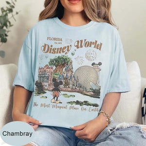 Może przedstawiać: Jasnoniebieska koszulka z grafiką Disney World na Florydzie. Grafika zawiera Myszka Miki, zamek i tekst "Disney World" oraz "The Most Magical Place On Earth."