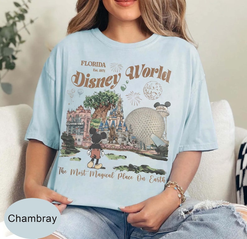 Może przedstawiać: Jasnoniebieska koszulka z grafiką Disney World na Florydzie. Grafika zawiera Myszka Miki, zamek i tekst "Disney World" oraz "The Most Magical Place On Earth."