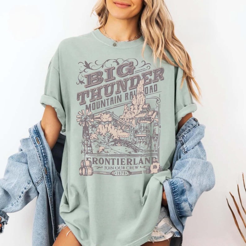 May include: A light sage green short-sleeve t-shirt featuring a dark brown graphic print of a mountain railroad scene. The text reads BIG THUNDER MOUNTAIN RAILROAD, FRONTIERLAND, JOIN OUR CREW, and 1979. The shirt is worn with a light blue denim jacket.