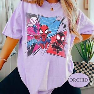 K&ouml;nnte beinhalten: Helllila T-Shirt mit einem Comic-Motiv von Spider-Man, Spider-Gwen und Miles Morales. Das Design zeigt kr&auml;ftige Farben und Actionposen. Das T-Shirt hat kurze &Auml;rmel und eine lockere Passform. Das Wort "ORCHID" ist aufgedruckt.