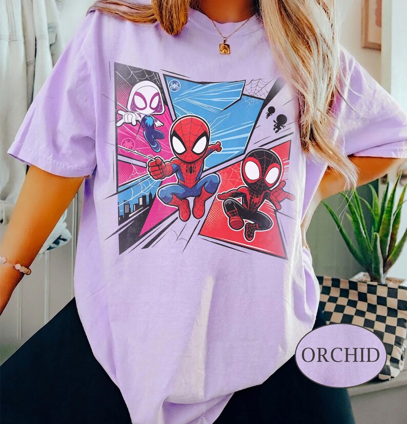 K&ouml;nnte beinhalten: Helllila T-Shirt mit einem Comic-Motiv von Spider-Man, Spider-Gwen und Miles Morales. Das Design zeigt kr&auml;ftige Farben und Actionposen. Das T-Shirt hat kurze &Auml;rmel und eine lockere Passform. Das Wort "ORCHID" ist aufgedruckt.