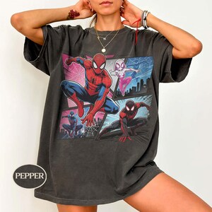 K&ouml;nnte beinhalten: Dunkelgraues T-Shirt mit einer Comic-Grafik von Spider-Man und anderen Spider-Verse-Charakteren. Das Design zeigt Spider-Man in Rot und Blau, Spider-Gwen in Pink und Wei&szlig; sowie Miles Morales in Schwarz und Rot. Das T-Shirt hat einen Vintage-Look.