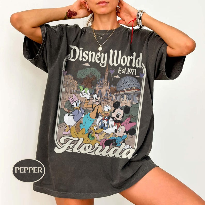 May include: Dark gray t-shirt featuring a Disney World graphic with Mickey Mouse, Minnie Mouse, Donald Duck, Daisy Duck, Goofy, and Pluto. The text "Disney World Est. 1971" and "Florida" are also displayed.