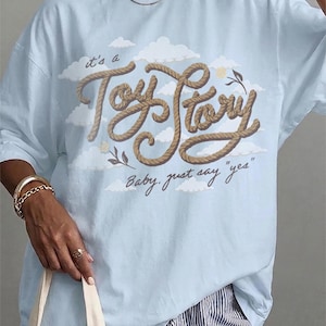 Pu&ograve; includere: Una t-shirt azzurro chiaro con una stampa grafica di nuvole bianche e il testo "it's a Toy Story" con texture a corda marrone sopra il testo in corsivo "Baby, just say "yes"" in marrone scuro, con piccoli accenti floreali ai lati.