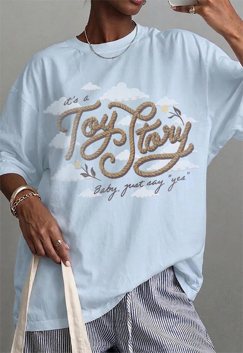 Pu&ograve; includere: Una t-shirt azzurro chiaro con una stampa grafica di nuvole bianche e il testo "it's a Toy Story" con texture a corda marrone sopra il testo in corsivo "Baby, just say "yes"" in marrone scuro, con piccoli accenti floreali ai lati.