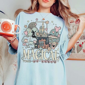 Puede incluir: Camiseta azul claro con un gr&aacute;fico colorido que presenta iconos de los parques de Disney, incluyendo un castillo, tazas de t&eacute; y un monorra&iacute;l. El texto dice "The Most Magical Place on Earth". En la imagen se sostiene una taza blanca con un dise&ntilde;o de sol.