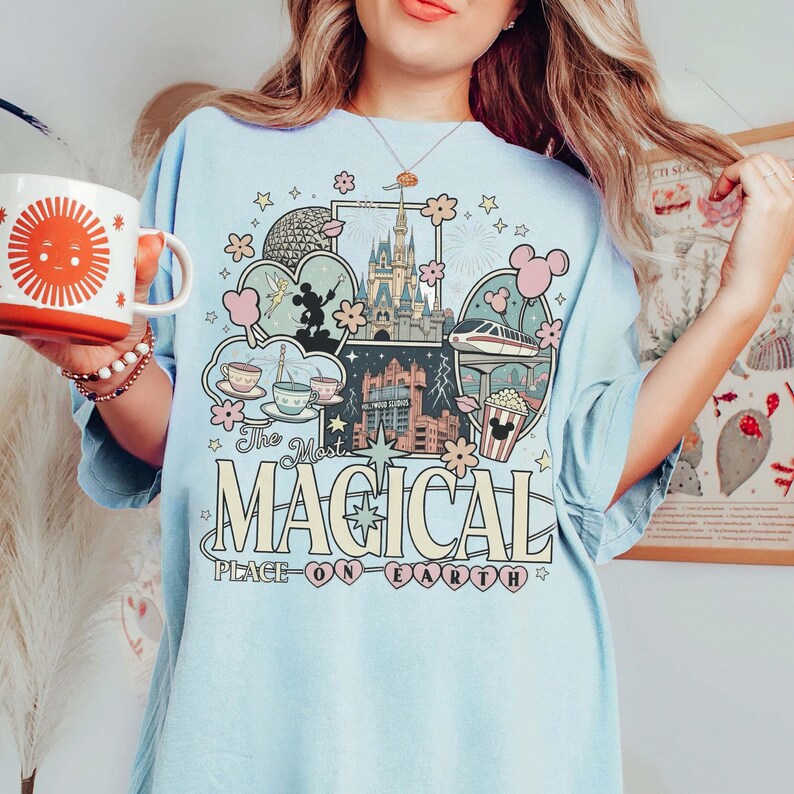 Puede incluir: Camiseta azul claro con un gr&aacute;fico colorido que presenta iconos de los parques de Disney, incluyendo un castillo, tazas de t&eacute; y un monorra&iacute;l. El texto dice "The Most Magical Place on Earth". En la imagen se sostiene una taza blanca con un dise&ntilde;o de sol.