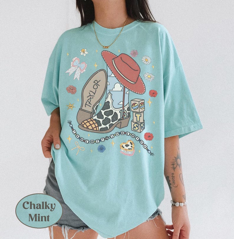 Puede incluir: Una camiseta de manga corta en color verde menta tiza con un estampado gr&aacute;fico de una bota vaquera con manchas de vaca, un sombrero rojo, un lazo, flores y bloques apilados con el texto Taylor, 13 y TS. Un dise&ntilde;o de cuentas enmarca el gr&aacute;fico.