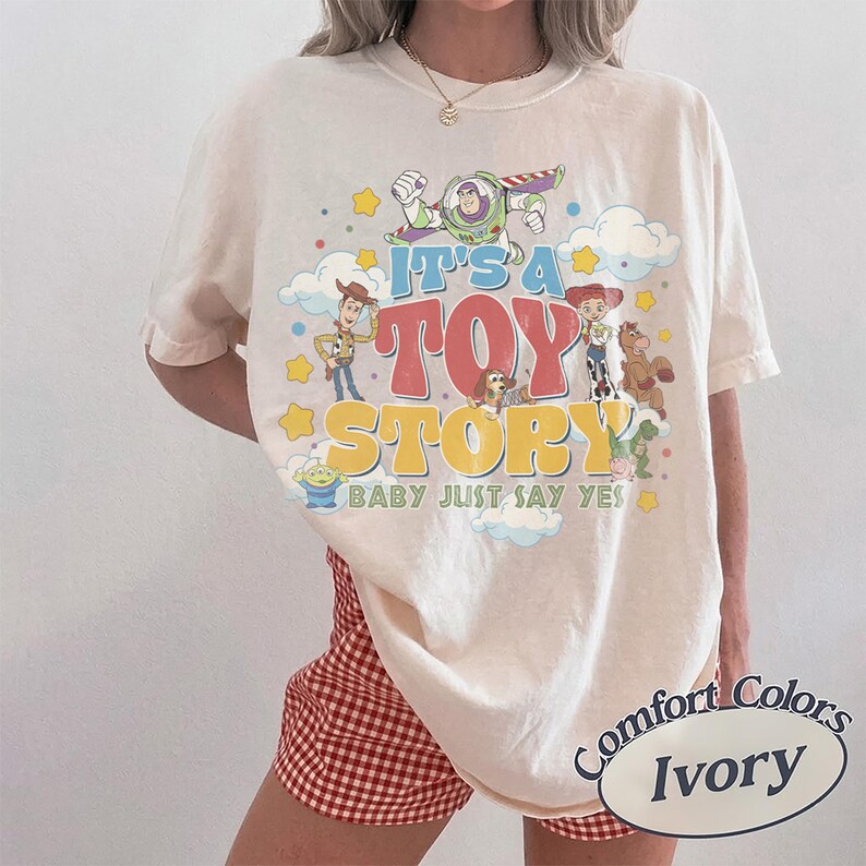 May include: An ivory short-sleeve t-shirt featuring a colorful graphic print of Toy Story characters including Woody, Buzz Lightyear, Jessie, and others. The text reads IT'S A TOY STORY BABY JUST SAY YES, surrounded by yellow stars and blue clouds.