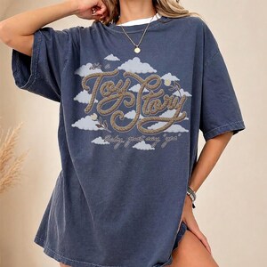 Pu&ograve; includere: Una t-shirt a maniche corte blu scuro con una stampa grafica di nuvole bianche e il testo "It's a Toy Story" scritto in un carattere marrone stile corda, con il testo aggiuntivo "Baby, just say 'yea'" stampato sotto in corsivo.