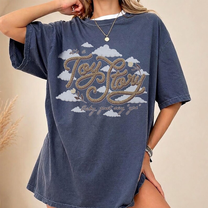 Pu&ograve; includere: Una t-shirt a maniche corte blu scuro con una stampa grafica di nuvole bianche e il testo "It's a Toy Story" scritto in un carattere marrone stile corda, con il testo aggiuntivo "Baby, just say 'yea'" stampato sotto in corsivo.