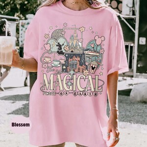 Puede incluir: Camiseta rosa claro con un dise&ntilde;o gr&aacute;fico colorido que presenta un castillo, tazas de t&eacute; y un monorra&iacute;l. El texto de la camiseta dice "The Most Magical Place on Earth". La camiseta es de corte holgado.