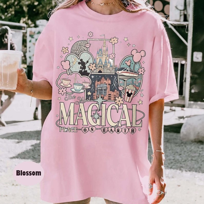 Puede incluir: Camiseta rosa claro con un dise&ntilde;o gr&aacute;fico colorido que presenta un castillo, tazas de t&eacute; y un monorra&iacute;l. El texto de la camiseta dice "The Most Magical Place on Earth". La camiseta es de corte holgado.