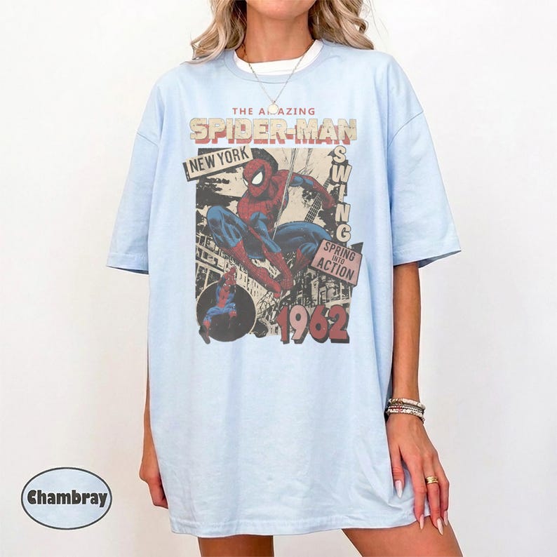 May include: A light blue t-shirt featuring a vintage-style graphic of Spider-Man in a dynamic pose. The print includes the text THE AMAZING SPIDER-MAN, NEW YORK, SPRING INTO ACTION, and 1962. The design shows a cityscape background in muted tones.