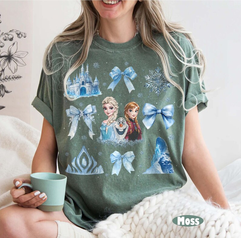 May include: A moss green t-shirt with a colourful graphic of characters and elements from a popular animated film. The design includes images of a castle, snowflakes, bows, and the characters Elsa, Anna, and Olaf.