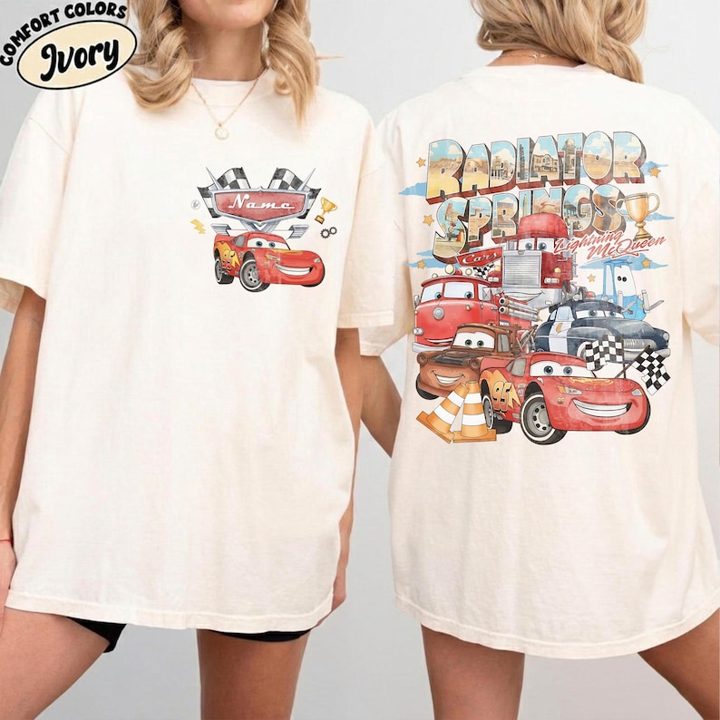 May include: Ivory t-shirt with a graphic of Lightning McQueen from the film Cars. The front shows the text "Name" and racing flags. The back of the shirt features "Radiator Springs" and other Cars characters in a vintage style.