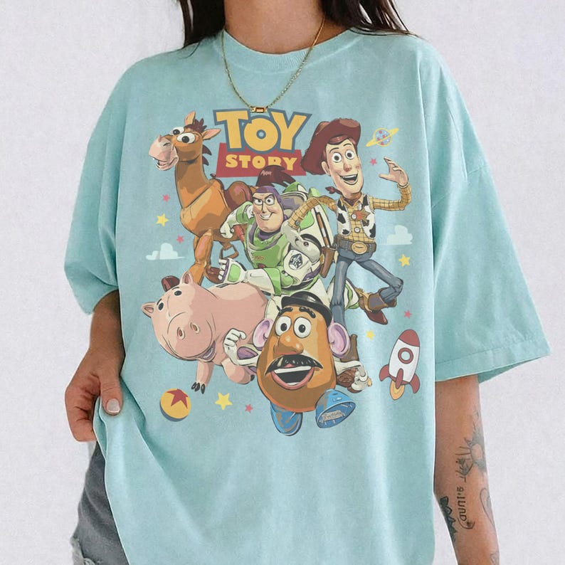 May include: A light blue t-shirt featuring a graphic print of Toy Story characters including Woody, Buzz Lightyear, Bullseye, Hamm, and Mr. Potato Head. The design includes the Toy Story logo, a small rocket, and colorful stars on the front.