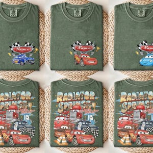May include: Green t-shirts with car-themed designs. Some shirts feature a race car design with a name banner, while others display the text "Radiator Springs" with various car illustrations. The shirts are displayed on woven mats.