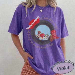May include: A violet short-sleeve t-shirt featuring a graphic of a tire framing cartoon characters, a red cowboy hat, and a red boot. The text reads Love has a way of bringing things back to life. The shirt has a crew neckline and a relaxed fit.
