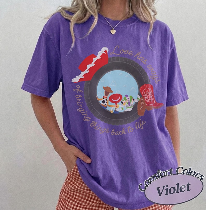 May include: A violet short-sleeve t-shirt featuring a graphic of a tire framing cartoon characters, a red cowboy hat, and a red boot. The text reads Love has a way of bringing things back to life. The shirt has a crew neckline and a relaxed fit.