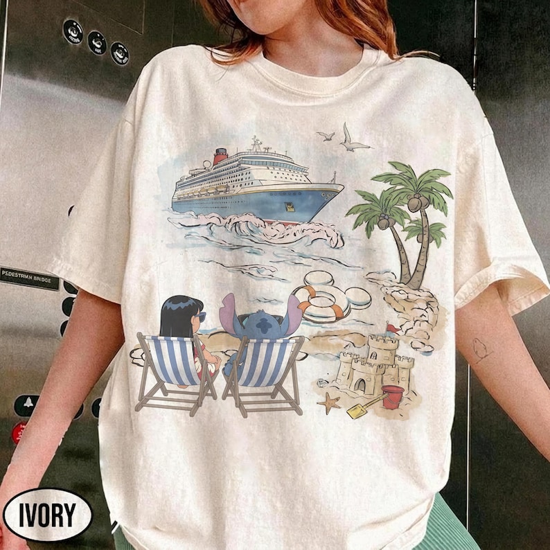 May include: Ivory-colored t-shirt featuring a beach scene with a cruise ship, palm trees, and cartoon characters. Two characters are in striped beach chairs. A sandcastle, starfish, and life preservers are also visible. The word "IVORY" is printed on the bottom left.