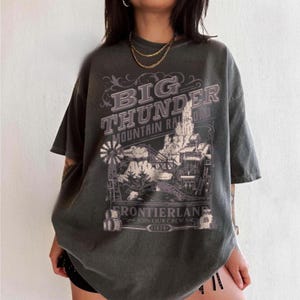 May include: A charcoal gray short-sleeve t-shirt featuring a graphic print with the text BIG THUNDER MOUNTAIN RAILROAD FRONTIERLAND JOIN OUR CREW 1979. The design includes a detailed illustration of a mountain landscape, a windmill, and barrels.