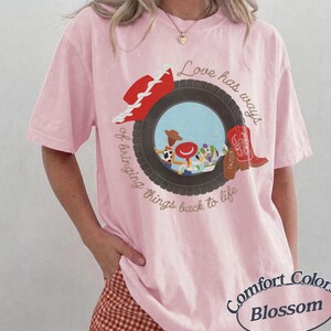 May include: A light pink short-sleeve t-shirt featuring a graphic of a black tire framing cartoon characters. The design includes a red hat, a red cowboy boot with the name Taylor, and the text Love has ways of bringing things back to life.